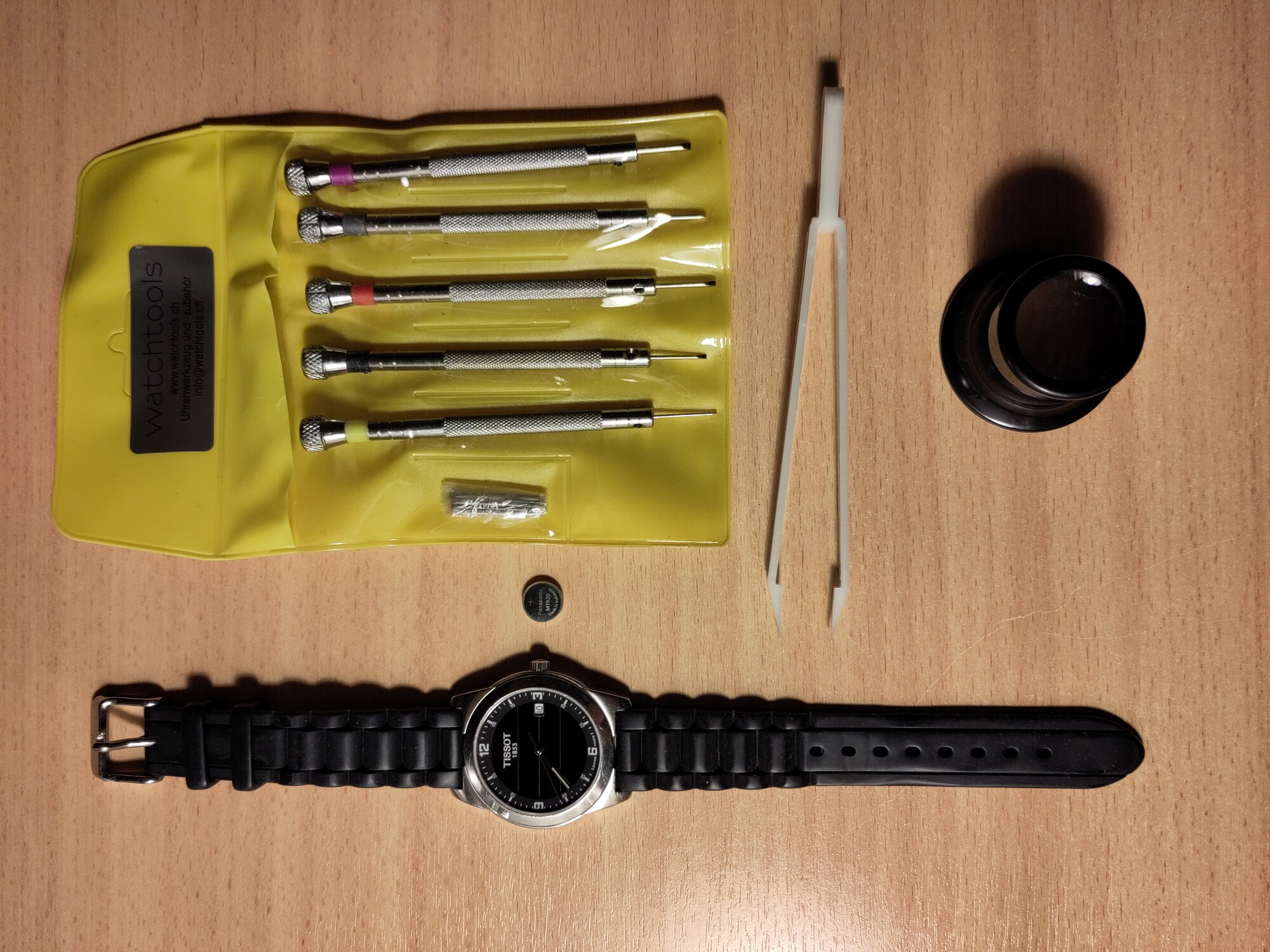 Eine Armbanduhr mit analogen Zeigern, daneben ein Akku in Knopfzellengrösse, Uhrmacherschraubenzieher, eine Pinzette und eine Uhrmacherlupe